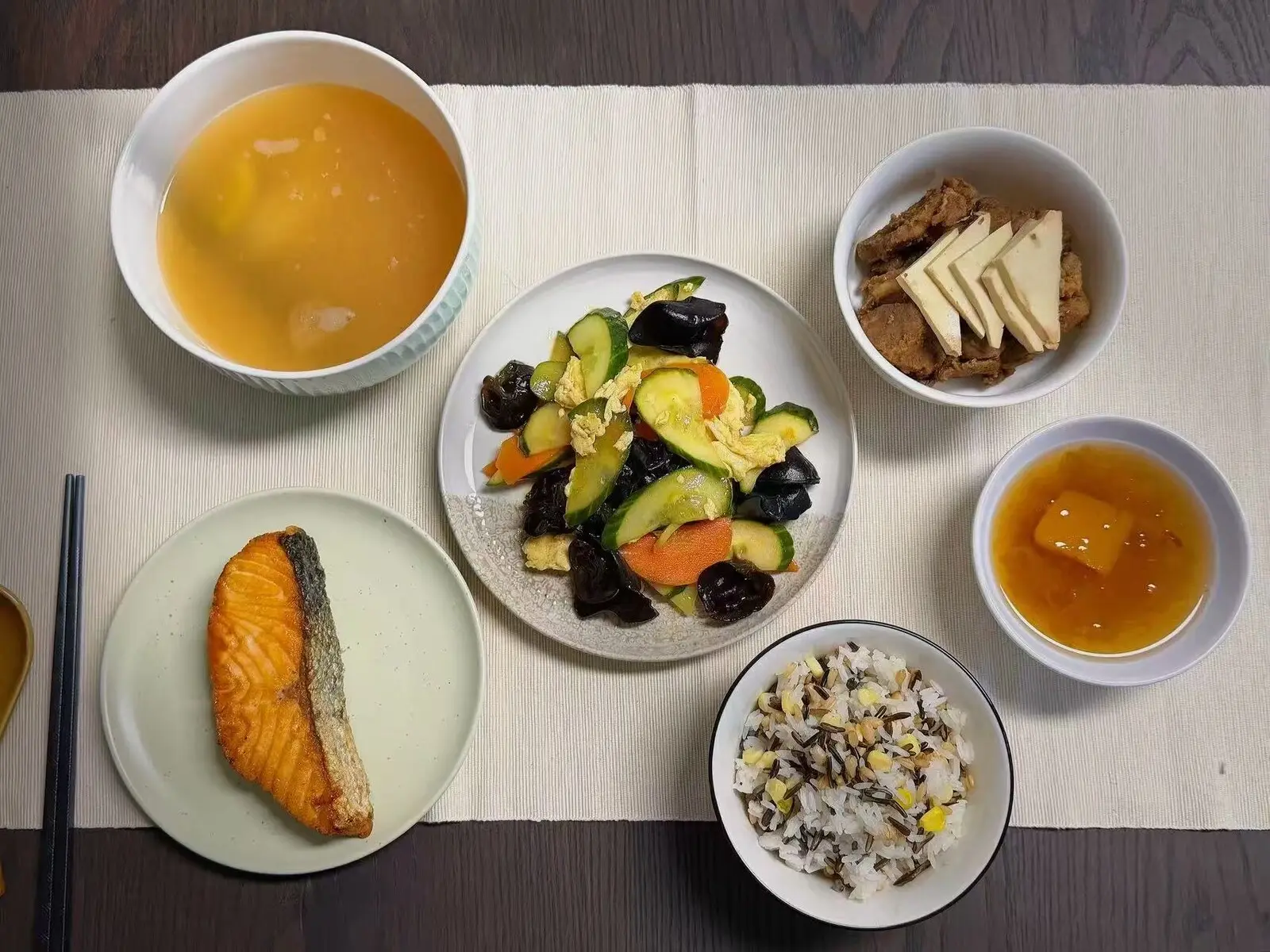 A Cantonese confinement meal seen from above on a cream table runner, with a bowl of soup, a pan-seared salmon fillet, a stir-fry of sliced gourd, wood ear mushrooms, carrot and egg, a bowl of braised meat with tofu, mixed-grain rice and a small bowl of amber broth