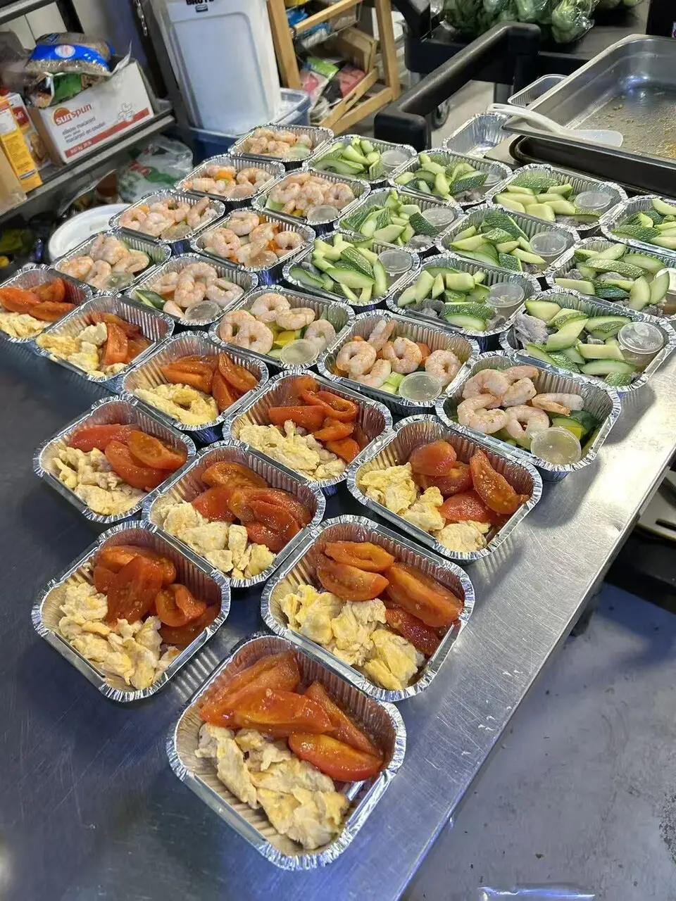 Rows of foil trays on a stainless-steel kitchen counter, filled with shrimp, sliced cucumber and tomato with scrambled egg, waiting to be portioned into meals