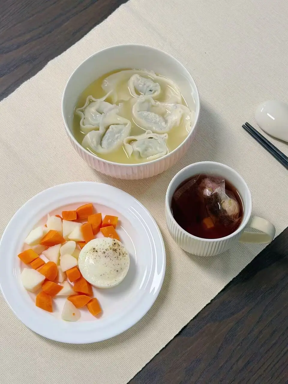 A bowl of dumplings in clear broth beside a plate of diced carrot and fruit with an egg, and a cup of hot tea with the tea bag still in it
