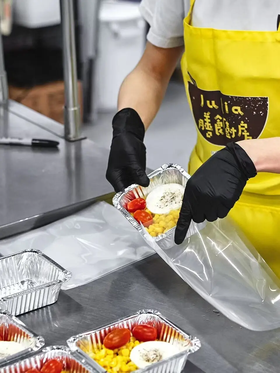 A kitchen worker in a yellow Julia's Kitchen apron and black gloves sliding a foil meal tray of egg, corn and cherry tomatoes into a clear bag