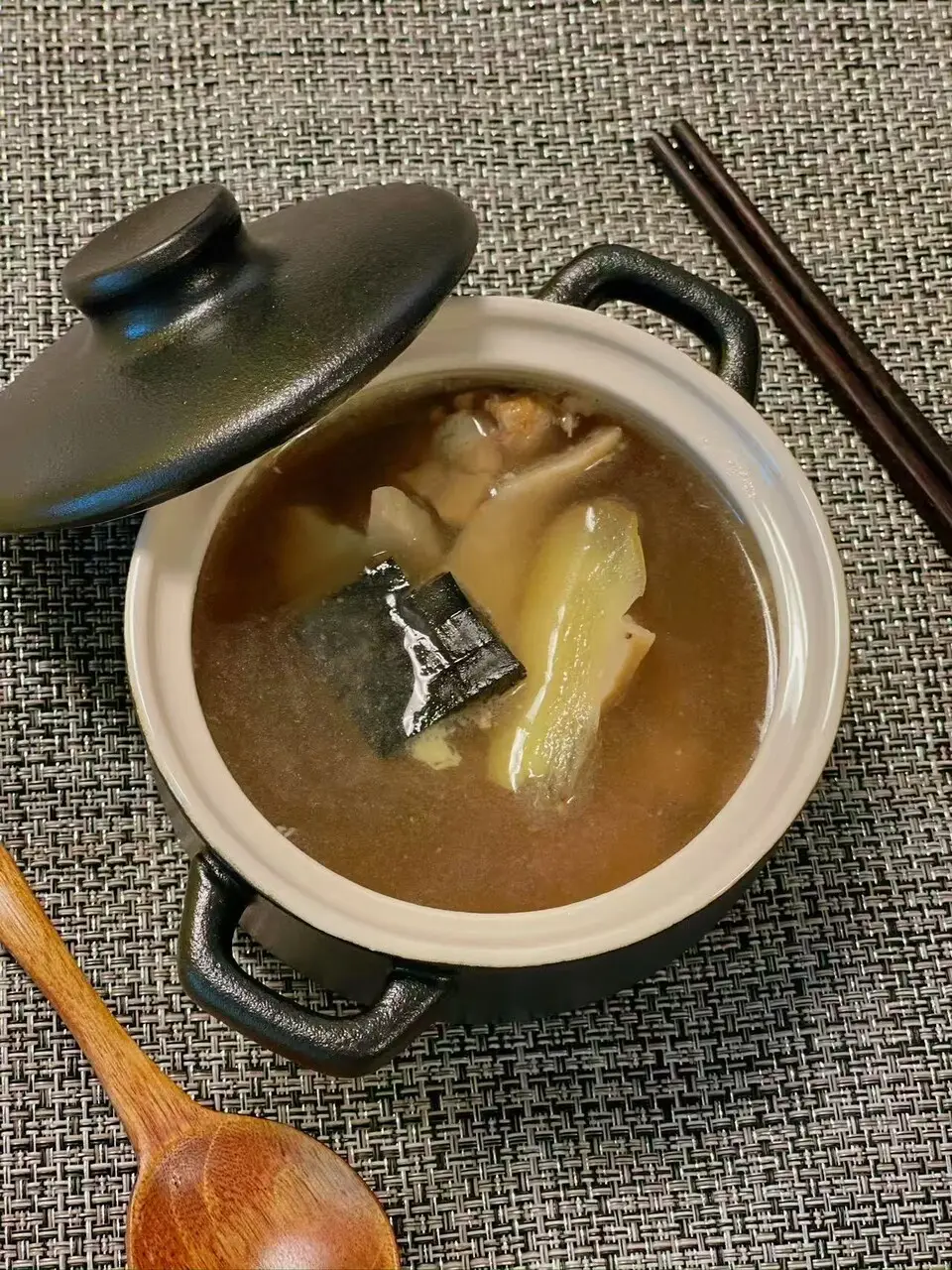 A small black claypot with its lid lifted, showing a slow-simmered soup in clear broth, with a wooden spoon and chopsticks beside it