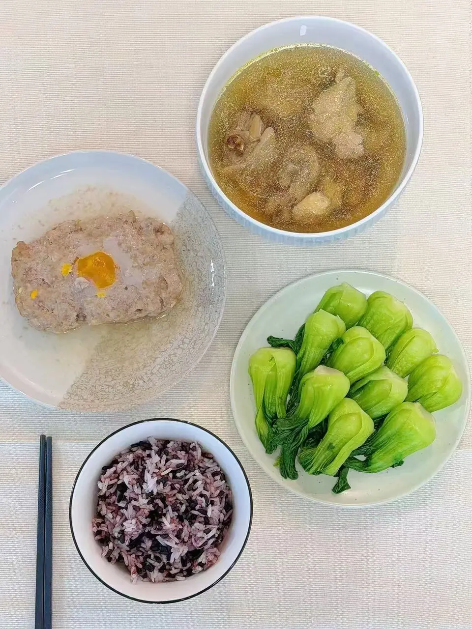 A confinement meal of steamed minced pork with a salted egg yolk, a bowl of chicken soup, a plate of Shanghai bok choy and a bowl of purple rice