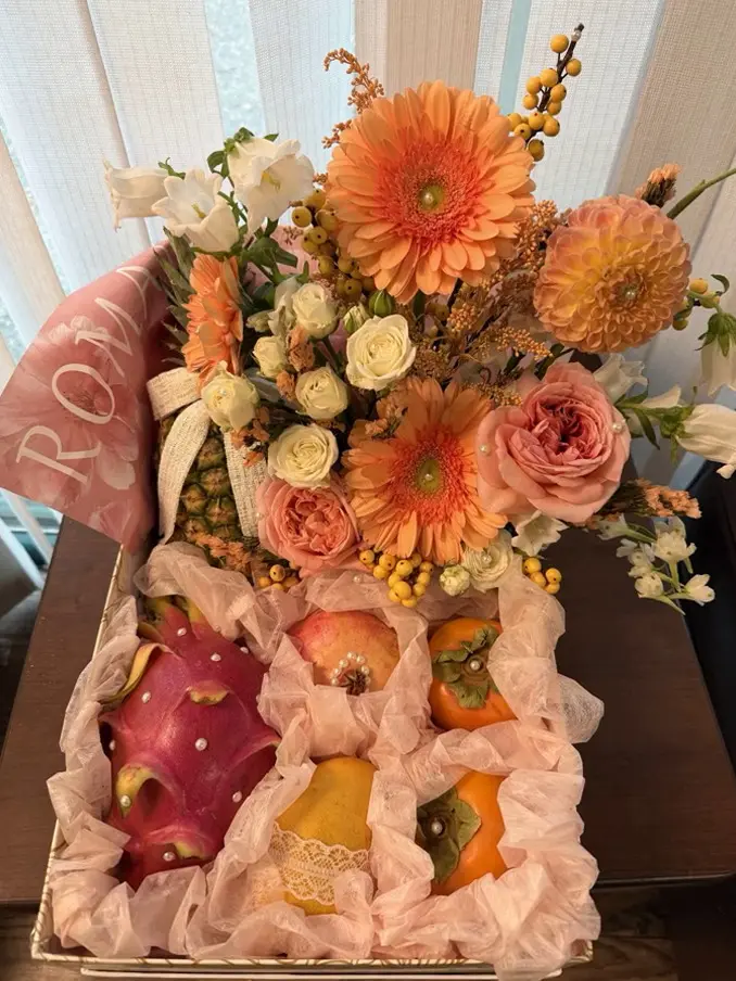 A gift basket for a mother, orange gerberas, a dahlia, peach and cream roses and sprigs of orange berries above a tray of fruit nested in pink tissue, with a dragon fruit, a pomegranate and two persimmons