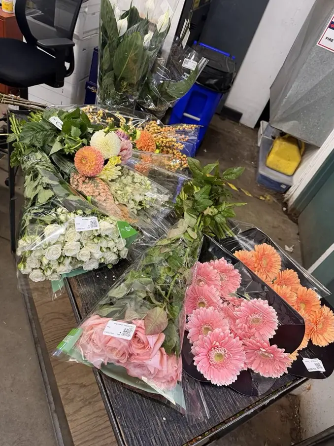 Freshly chosen flowers in clear cellophane on a cart, pink and orange gerberas, pink roses, white spray roses and greenery, ready to be arranged into gift baskets