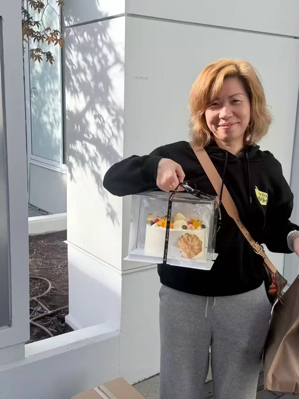 Julia, in a black hoodie with the Julia's Kitchen logo, smiles outside a client's building as she carries a fresh-fruit cream cake in a clear gift box for a mother who has just given birth