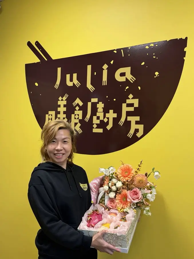 Julia, in a black hoodie with the Julia's Kitchen logo on the chest, smiles in front of the yellow Julia's Kitchen logo wall, holding a gift basket with orange and pink gerberas and roses on top and fresh fruit below