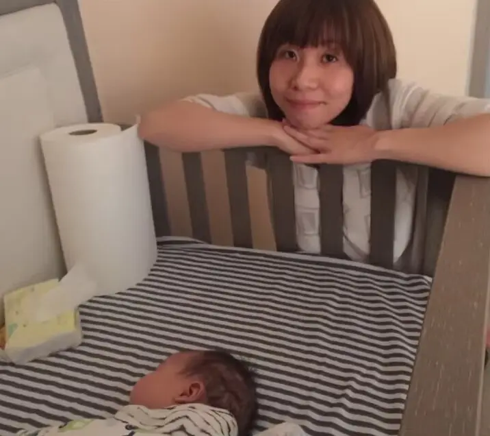 Julia smiling at the camera with her chin resting on her folded hands on the rail of a wooden crib, where a newborn sleeps on a grey-and-white striped sheet