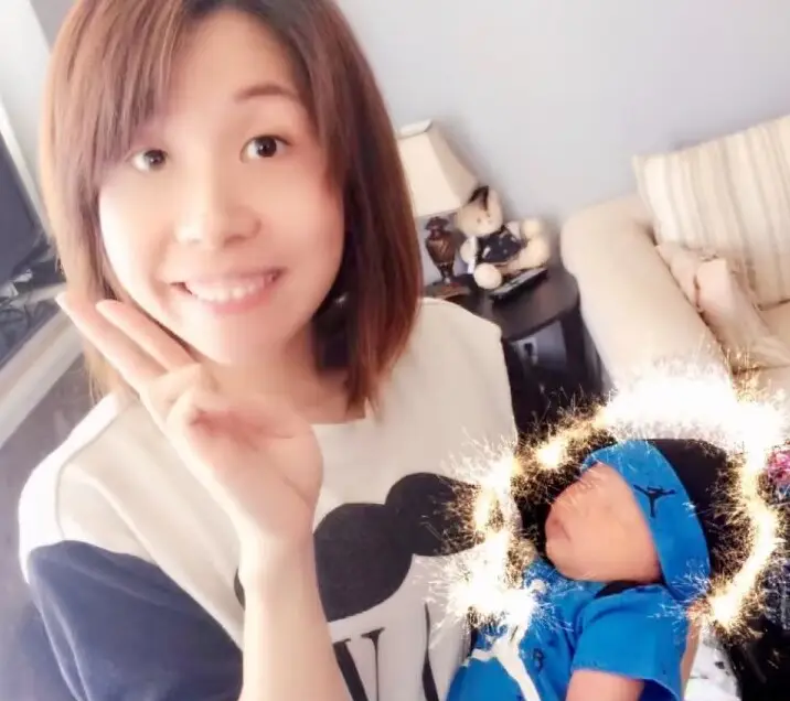 Julia taking a cheerful selfie in a living room, making a hand gesture, beside a sleeping baby in a blue cap and blue outfit
