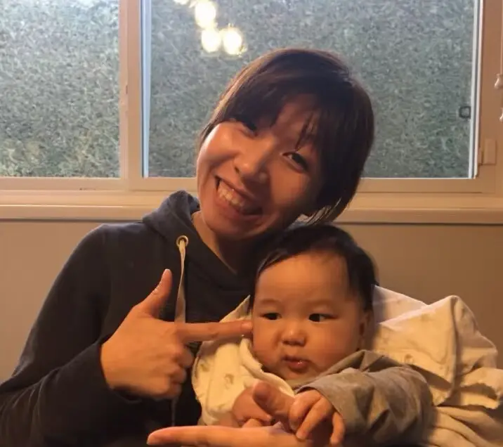 Julia sitting by a window with a baby in her arms, smiling and pointing to coax the baby to look at the camera