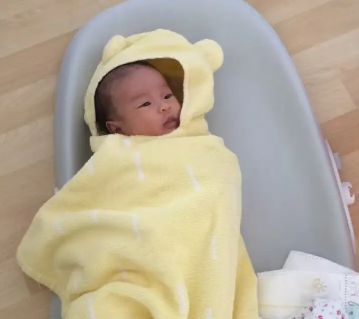 A newborn wrapped in a yellow hooded towel with little bear ears, lying awake in a grey baby bath support