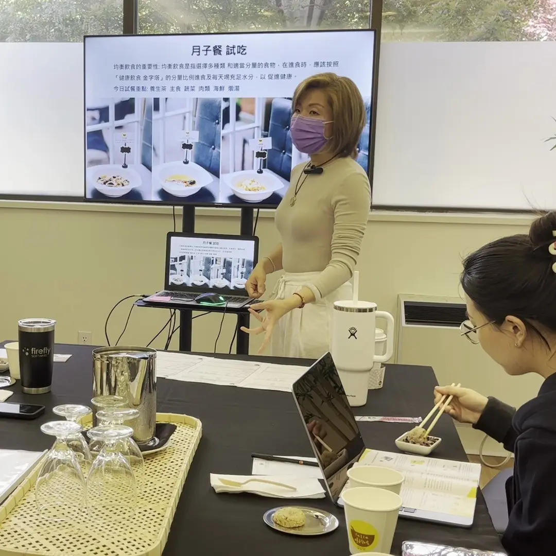 Julia presenting at a Cantonese confinement meal tasting session in Vancouver, beside a slide headed 月子餐 試吃 that sets out the day's tasting line-up of herbal tea, rice, vegetables, meat, seafood and slow-simmered soup, while a guest in the foreground tastes a sample with chopsticks